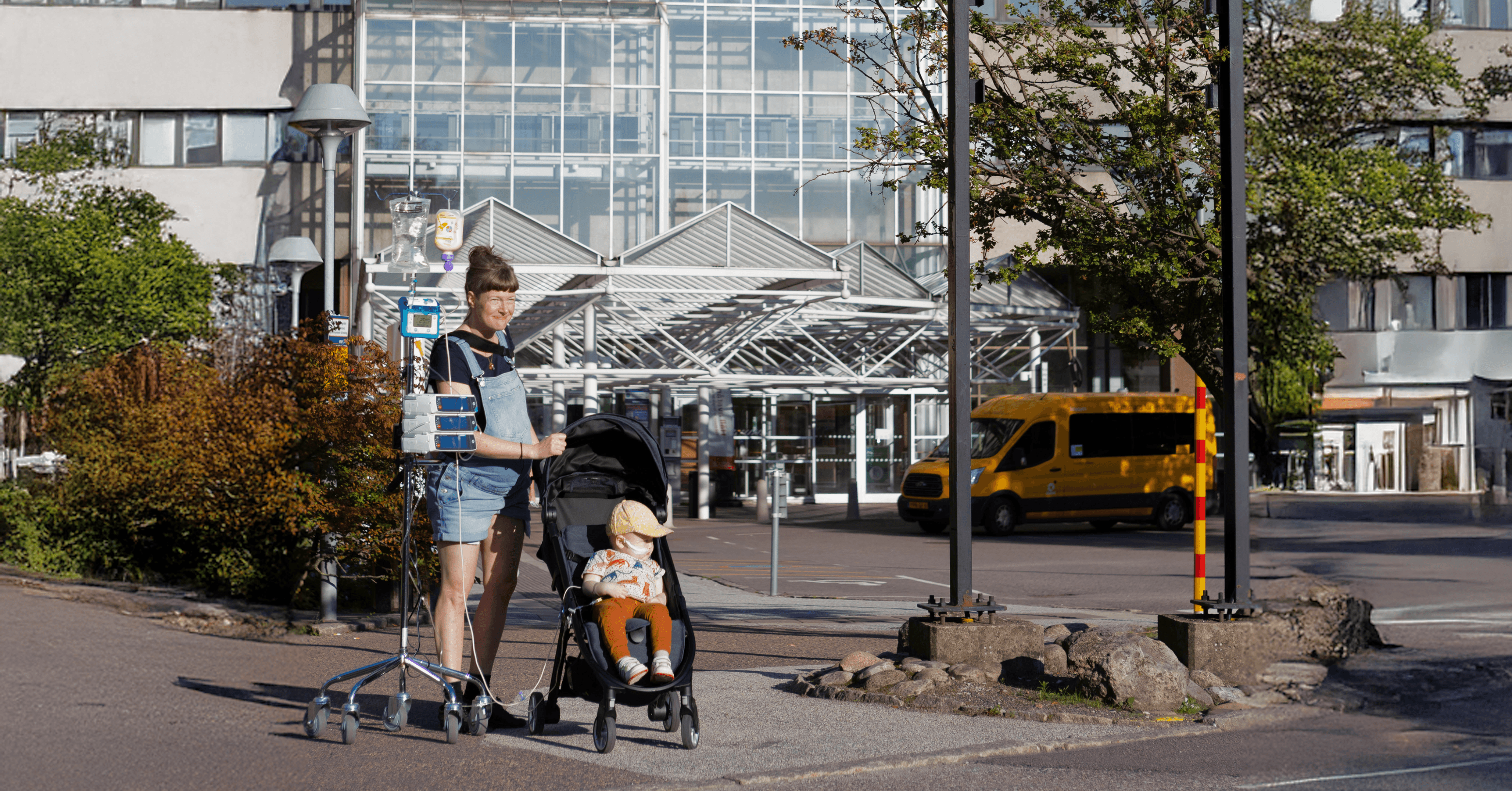 A person is pushing a child in a stroller outside a building, accompanied by an IV stand.