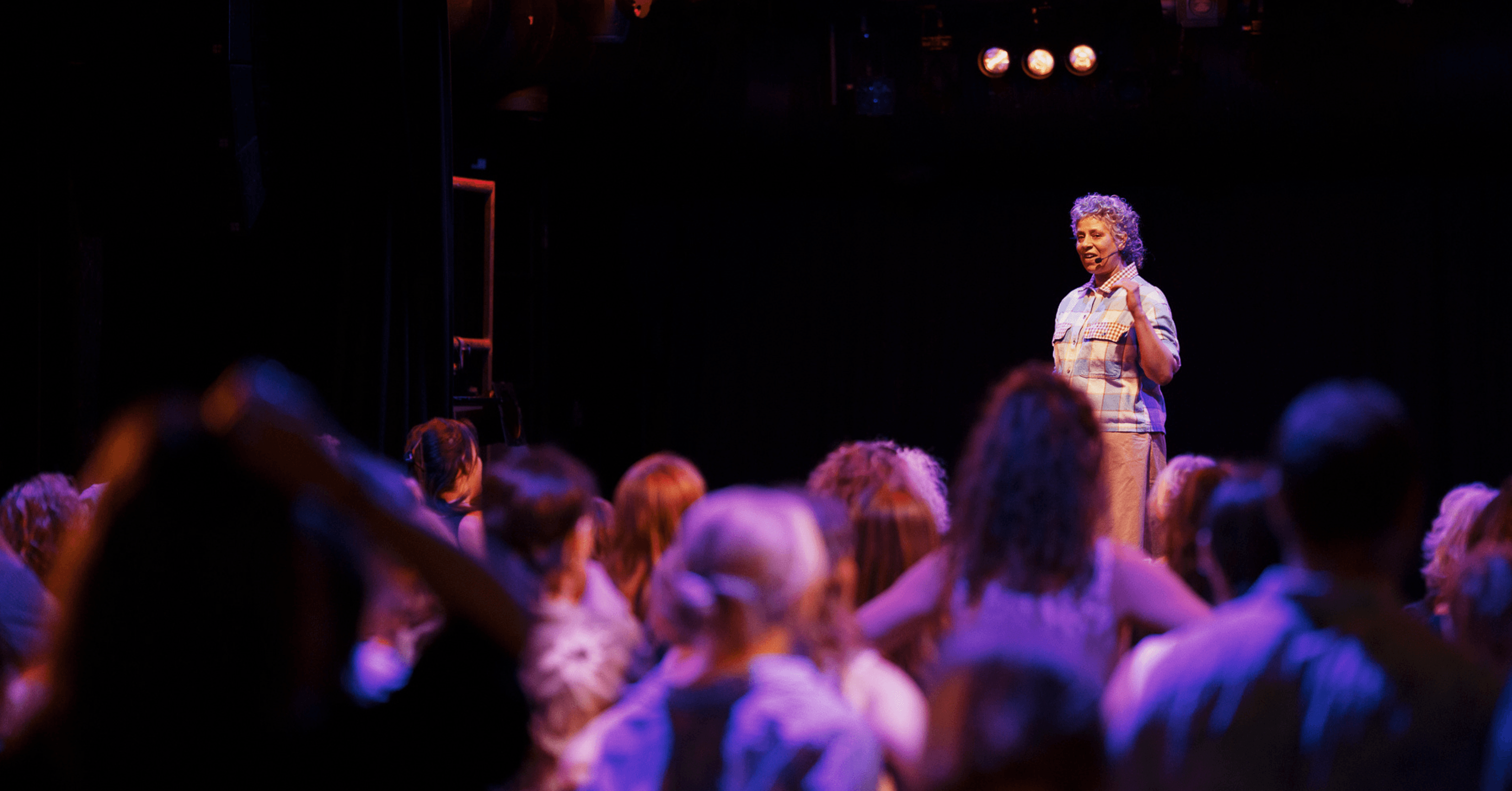 A person is speaking on stage to an audience in a dimly lit setting with colorful lighting.